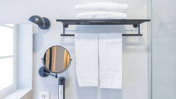 Un baño con cabina de ducha de vidrio, un perchero negro con toallas dobladas, un espejo redondo con marco negro y una lámpara fijada a la pared.