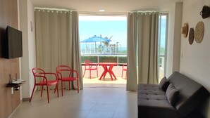 A living room with a grey sofa, red chairs, a TV mounted on the wall, and a balcony with a table and chairs. - Frente Mar com Terraço Privativo e Pet Friendly (Barra de São Miguel)