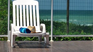 A white lounge chair with a blue towel and a straw hat on it, positioned on a wooden deck with a view of the ocean through a wire mesh fence. - Frente Mar com Terraço Privativo e Pet Friendly (Barra de São Miguel)