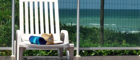 A white lounge chair with a blue towel and a straw hat on it, positioned on a wooden deck with a view of the ocean through a wire mesh fence.