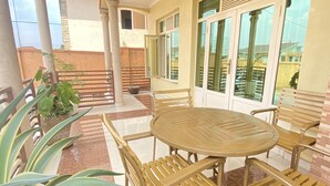 Een overdekt terras met een ronde tafel en stoelen, plantenbakken en uitzicht op de omliggende gebouwen.