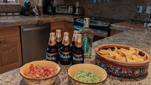 A kitchen with granite countertops, wooden cabinets, and a microwave. There are bowls of guacamole and salsa, a bowl of tortilla chips, and several bottles of Modelo beer.