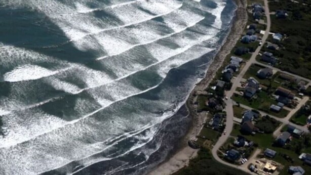 Vista aérea de una zona residencial costera con casas a lo largo de la orilla, una carretera curva y una pequeña playa.