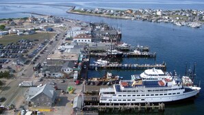 Un muelle de transbordador con una embarcación blanca grande, un estacionamiento y un pequeño edificio con un letrero.