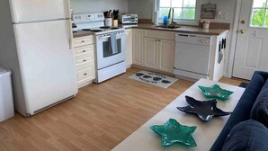 Cocina compacta con gabinetes blancos, refrigerador, microondas, estufa y lavavajillas. Hay una ventana que proporciona luz natural y una encimera con fregadero.