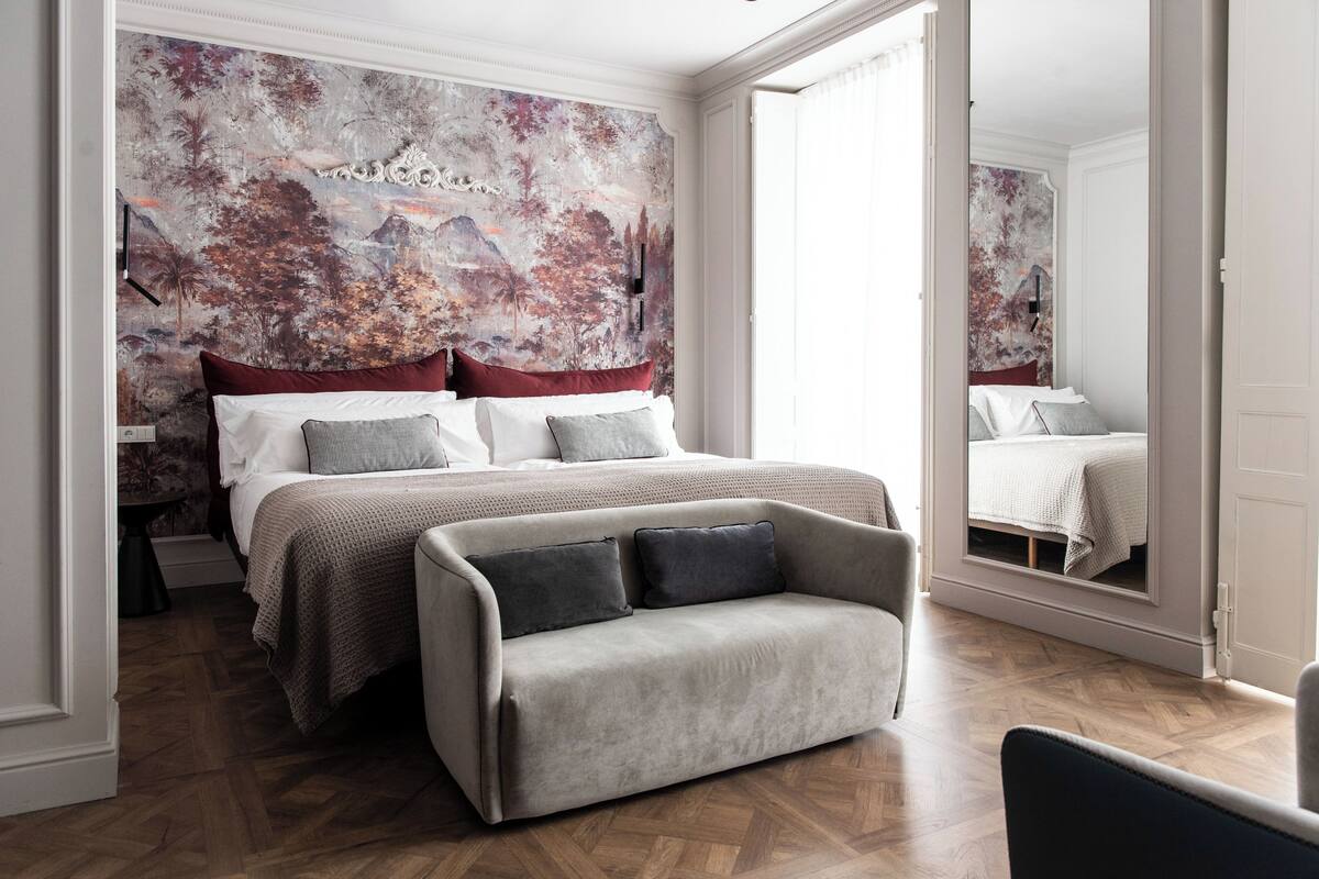 A bedroom with a large bed, a sofa, a mirror, and a mural on the wall.