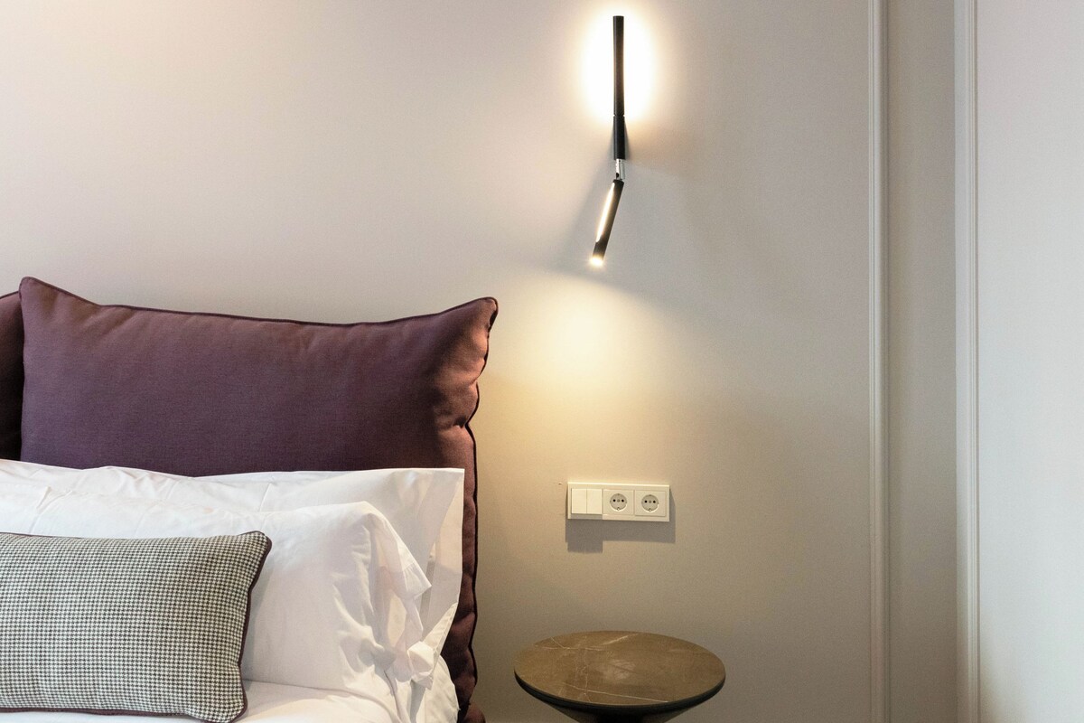 A neatly made bed with white and purple bedding, a small round table, and a wall-mounted light fixture.