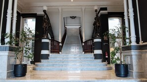 Una imponente escalera con escalones de mármol y barandillas negras, flanqueada por columnas y plantas en macetas.