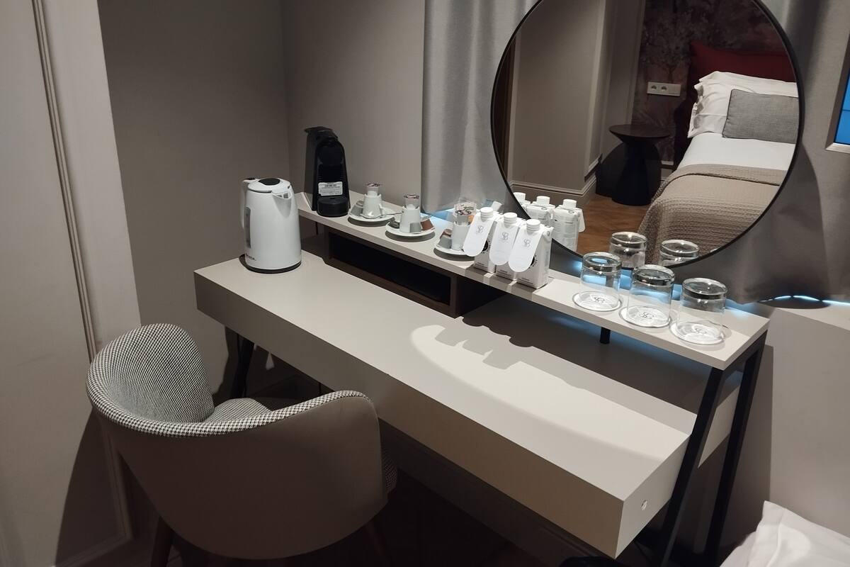 A hotel room with a round mirror, a white vanity table, a coffee maker, and toiletries.