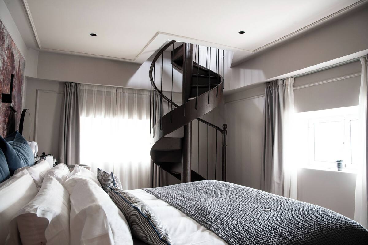 A bedroom with a modern design, featuring a spiral staircase, a neatly made bed, and a large window with curtains.