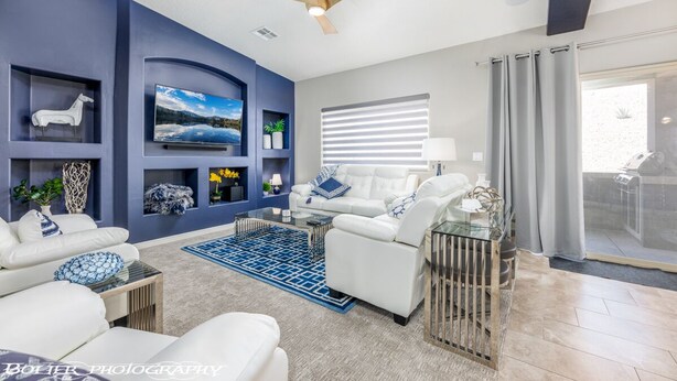 Uma sala de estar moderna com sofá branco, uma parede azul de destaque, ventilador de teto e uma janela grande com persianas.