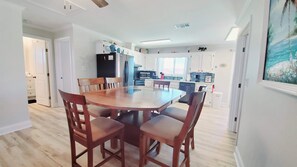 A dining area with a wooden table and chairs, a kitchen in the background, and a painting on the wall. - Salty Lime, Pet Friendly! Beach House!  Sleeps 8 Steps from the beach! (Gulf Shores)