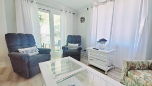 A living room with two dark armchairs, a white side table with a lamp, and a glass-top coffee table. - Salty Lime, Pet Friendly! Beach House!  Sleeps 8 Steps from the beach! (Gulf Shores)