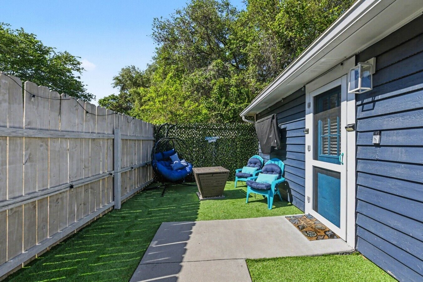 A backyard with artificial grass, a wooden fence, a blue hanging chair, and two turquoise chairs.