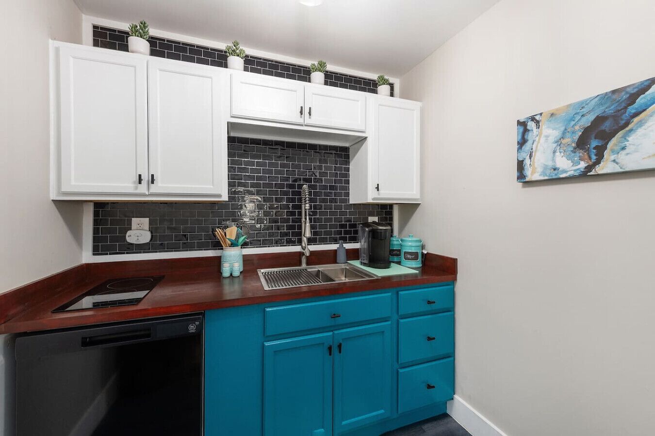 Cocina compacta con armarios superiores blancos, salpicadero de azulejos negros, encimera azul y lavavajillas.