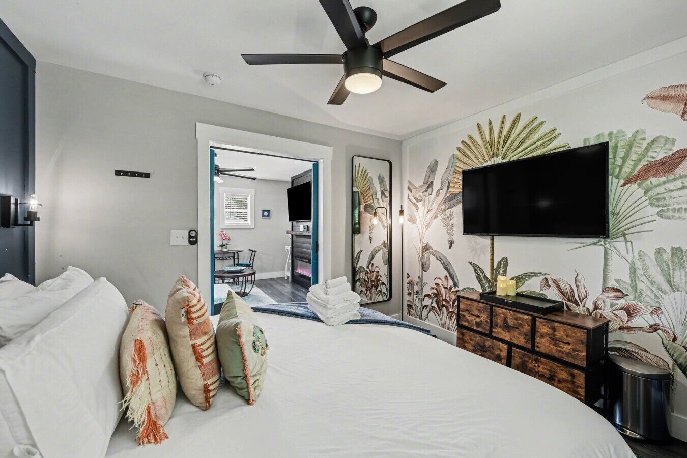 Une chambre à coucher avec un lit, des oreillers, une télévision, un ventilateur de plafond et un mur orné d’un motif floral.