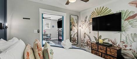 Une chambre à coucher avec un lit, des oreillers, une télévision, un ventilateur de plafond et un mur orné d’un motif floral.