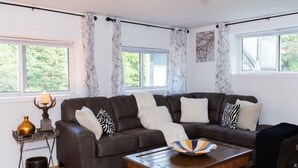 A living room with a brown leather sofa, a wooden coffee table, and a white rug. There are two windows with curtains, a lamp on a side table, and a framed picture on the wall. - Cozy Family Cottage, Sleeps up to 10 Persons & Fully Equipped (Maynooth)