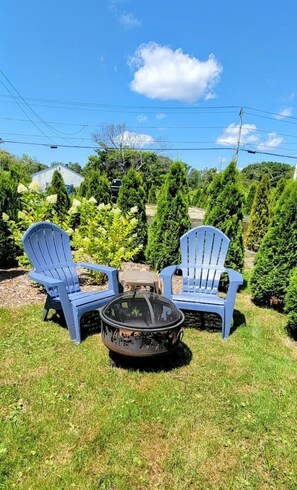 Zwei blaue Adirondack-Stühle und eine schwarze Feuerstelle in einer grünen Außenanlage mit Bäumen und einem Haus im Hintergrund.