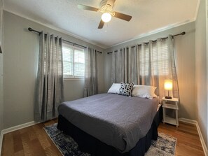 Ein Schlafzimmer mit Bett, Vorhängen, Deckenventilator, Nachttisch mit Lampe und Fenster mit Jalousien.