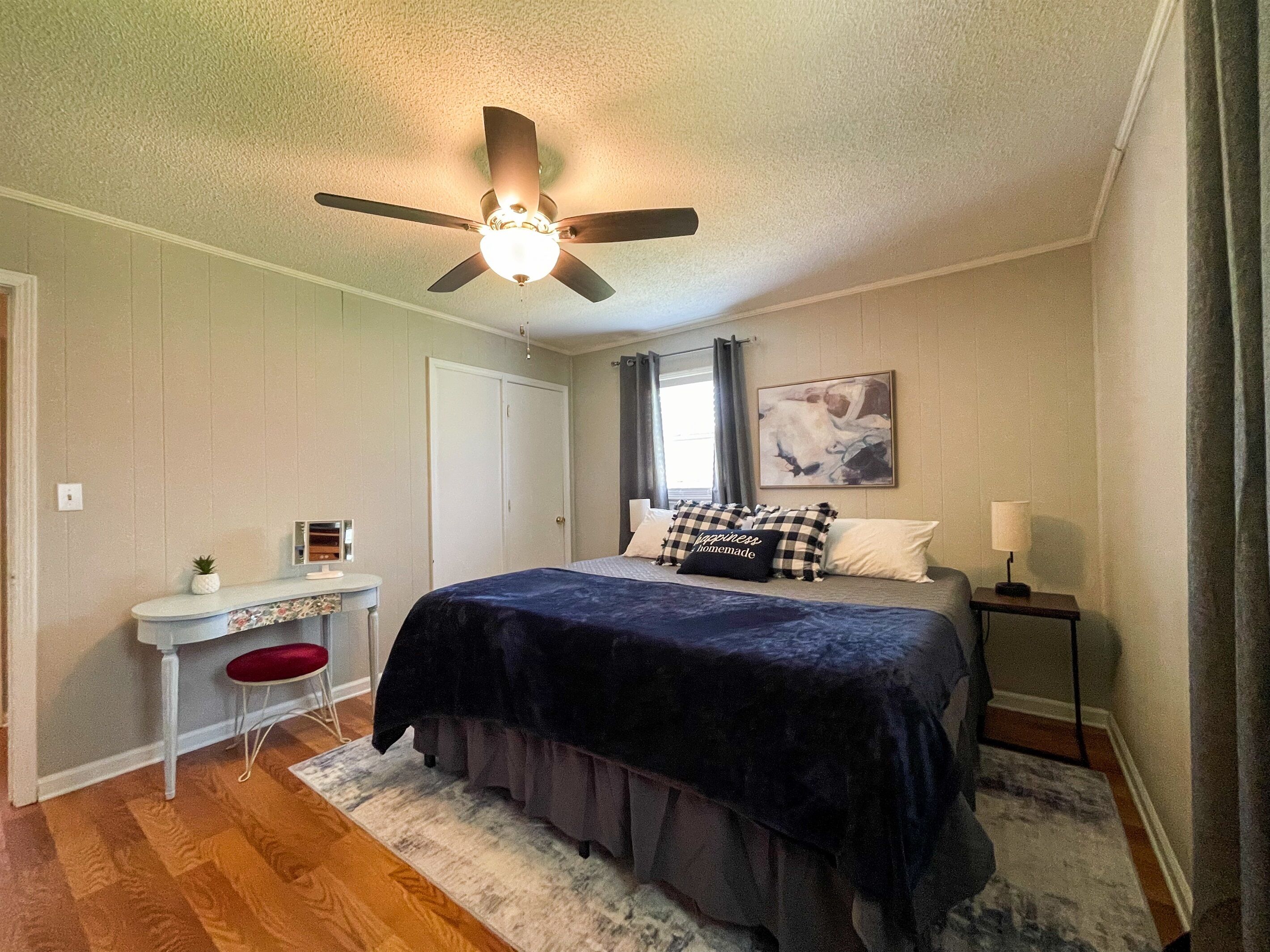 A bedroom with a bed, a desk, a chair, a ceiling fan, and a window with curtains.
