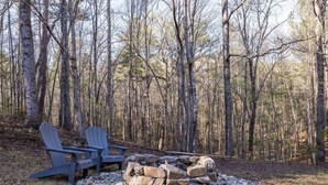Een bebost gebied met een vuurkorf van stenen en twee Adirondack stoelen.