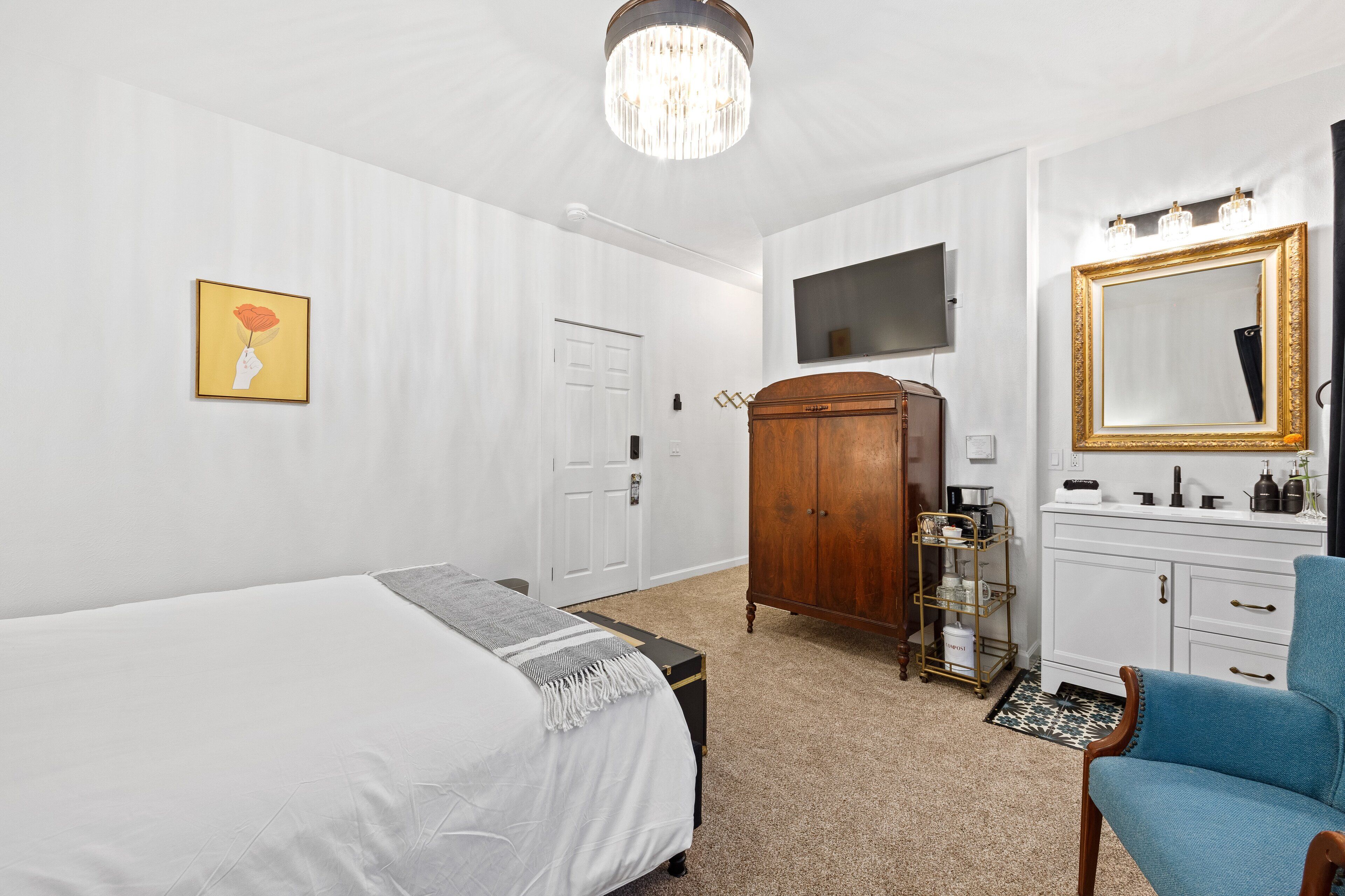 A hotel room with a bed, a wooden wardrobe, a vanity with a mirror, and a blue armchair.