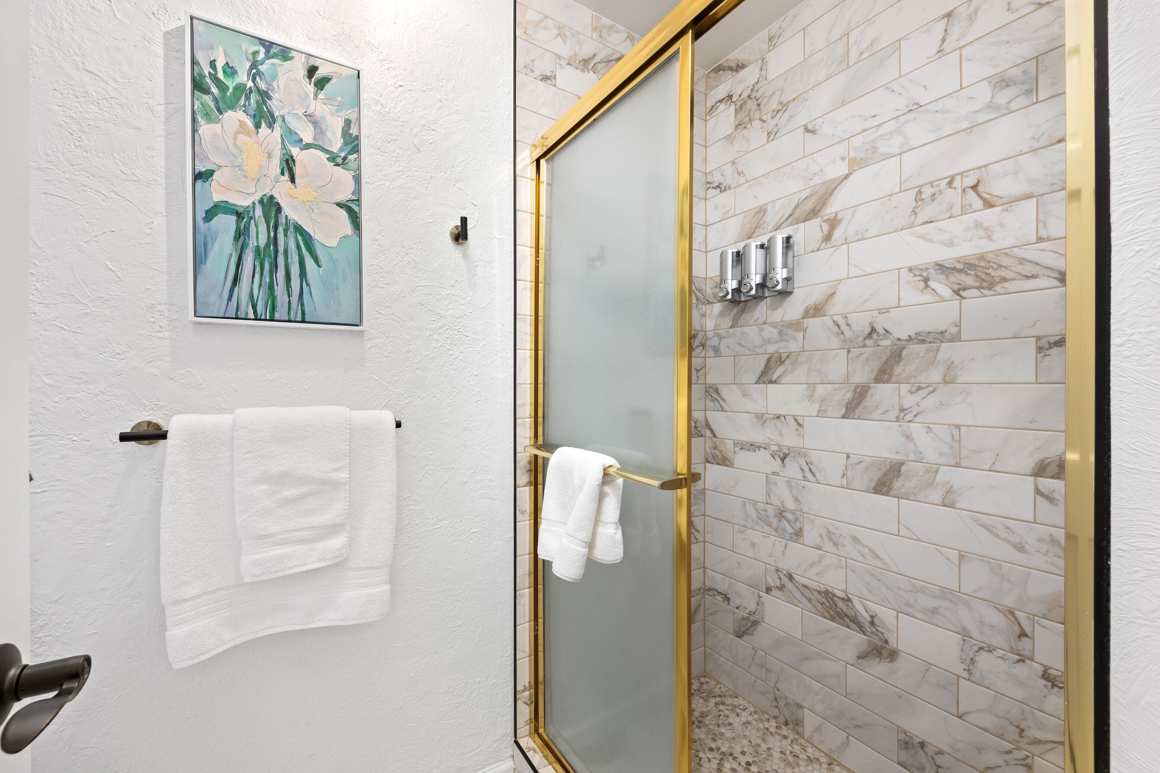 A bathroom with a glass shower door, marble tiles, and a painting of flowers on the wall.
