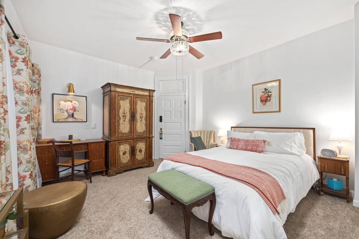 A bedroom with a bed, a desk, a chair, a cabinet, a ceiling fan, and a bedside table with a lamp.