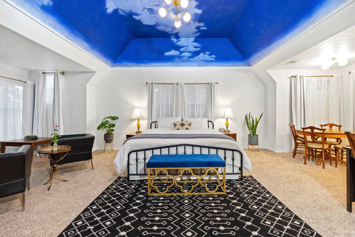 A bedroom with a starry sky ceiling, a bed with a blue headboard, a bedside table with a lamp, and a dining area with a table and chairs.