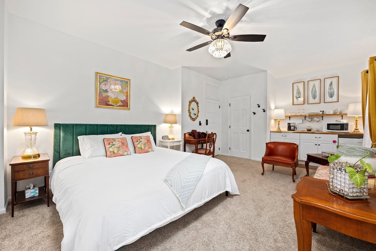 A hotel room with a large bed, a ceiling fan, a desk with a lamp, a chair, and a small table with a plant.