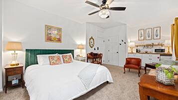 A hotel room with a large bed, a ceiling fan, a desk with a lamp, a chair, and a small table with a plant.