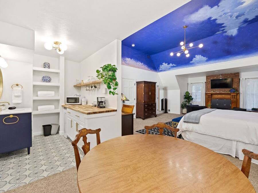 A modern hotel room with a dining area, a large bed, a TV, and a ceiling mural.