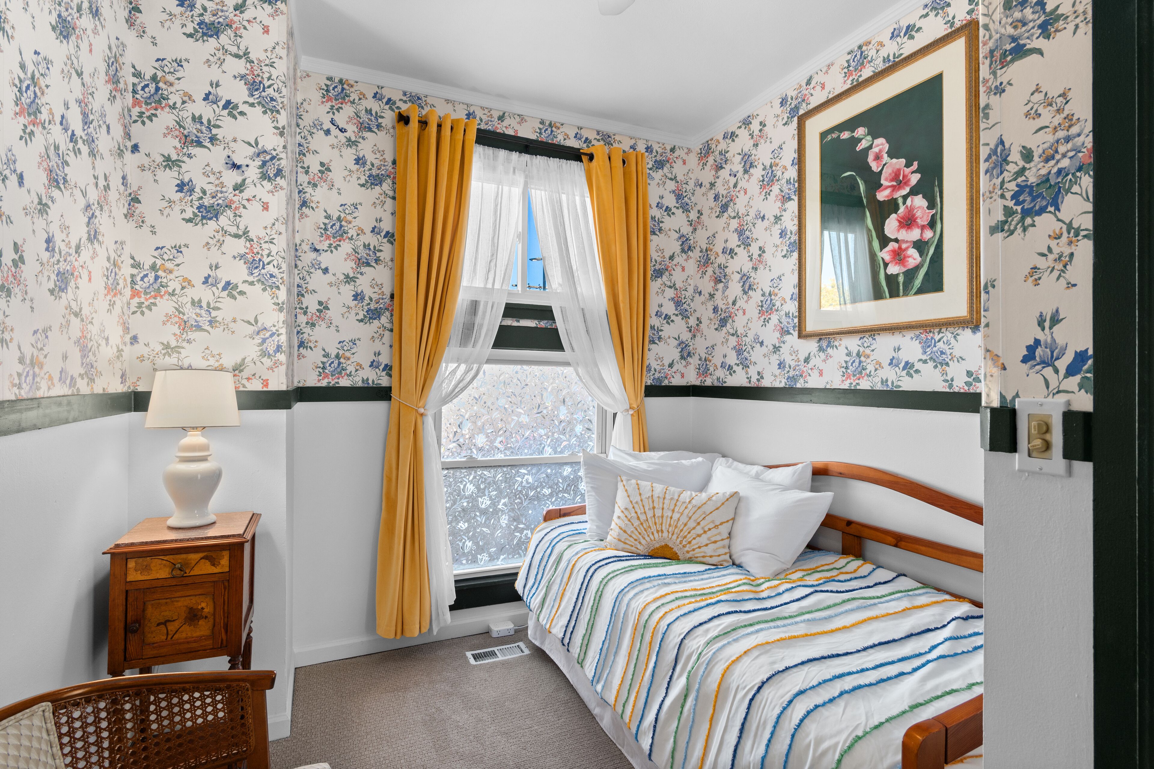 Un dormitorio con papel tapiz floral, una cama con ropa de cama a rayas, una mesita de noche con una lámpara y una ventana con cortinas amarillas.
