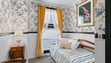 Un dormitorio con papel tapiz floral, una cama con ropa de cama a rayas, una mesita de noche con una lámpara y una ventana con cortinas amarillas.