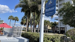 A sign for 'Beachhouse Suites & Condos' with a sailboat logo, surrounded by palm trees and a white building with balconies.