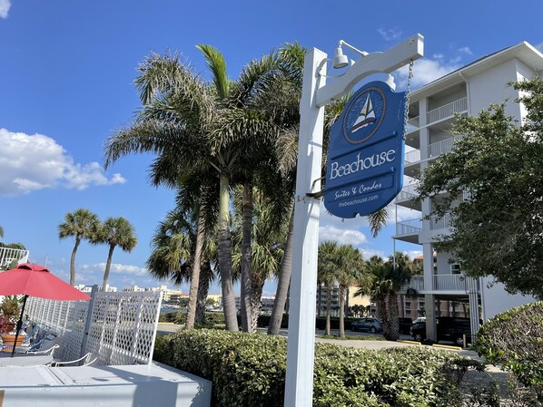A sign for 'Beachhouse Suites & Condos' with a sailboat logo, surrounded by palm trees and a white building with balconies.