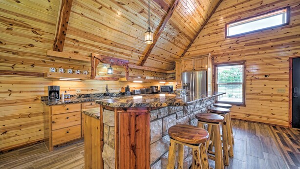 Interior de una cabaña de madera con cocina, que incluye encimera de granito, taburetes de bar y una isla de piedra.