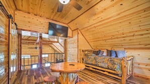 Interior de una cabaña de madera con una cama, un televisor instalado en el techo y una mesa de comedor con taburetes.