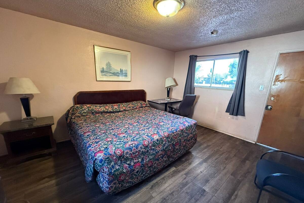 A small, neatly arranged room with a bed, a desk, a chair, a window with curtains, and a door.
