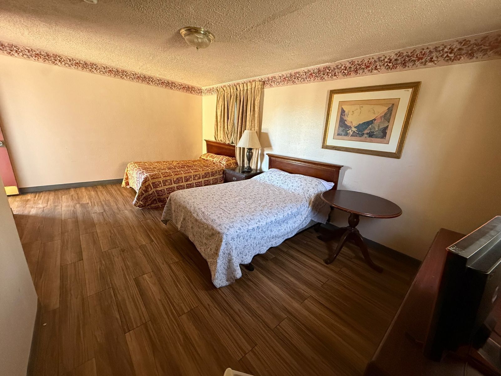 A hotel room with two beds, a small round table, a lamp, a painting on the wall, and a television.