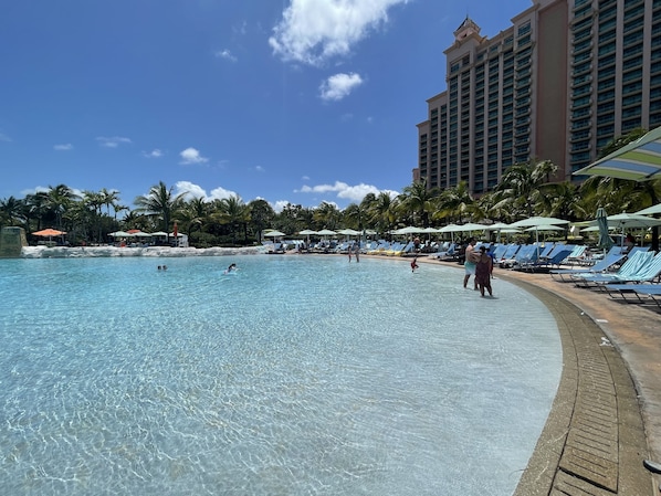 Una amplia piscina al aire libre con agua cristalina, rodeada de sillones de playa y sombrillas, con un alto edificio de hotel al fondo.