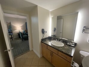 A hotel bathroom with a sink, mirror, and toilet. There is a small table with a lamp and a chair in the adjacent room. - 5 Br. 3 units. Sleeps 16  December 19-26, 2026 XMAS IN PARADISE BRING THE FAMILY (Nassau)