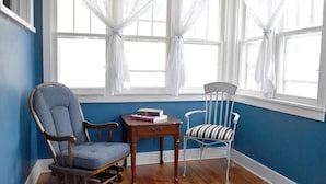 A room with a blue wall, wooden flooring, a rocking chair, a metal chair, a small table with books, and a window with white curtains. - Historic Bluebell. Entire House. 2Br Cozy but spacious (Wheatland)