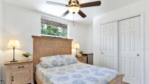 Une chambre à coucher comprenant un lit, deux tables de chevet avec des lampes, un ventilateur de plafond, une fenêtre et une porte double.