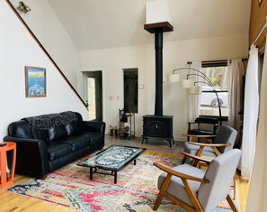 En stue med en sort sofa, et sofabord og to lænestole. Der er en sort brændeovn i hjørnet, og et vindue med gardiner.