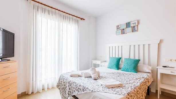 A neatly arranged bedroom with a bed, bedside tables, a TV, and a window with sheer curtains. - A&n Levante Torre del Mar (Torre del Mar)