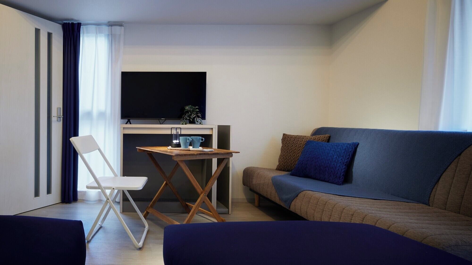 Un espacio habitable compacto con sofá, una mesita con una silla, televisor montado en la pared y ventana con cortinas.