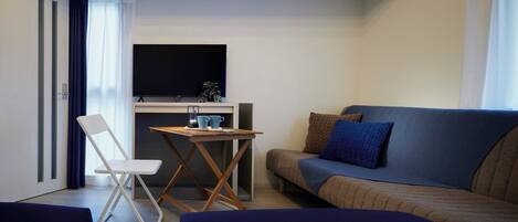 Un espacio habitable compacto con sofá, una mesita con una silla, televisor montado en la pared y ventana con cortinas.