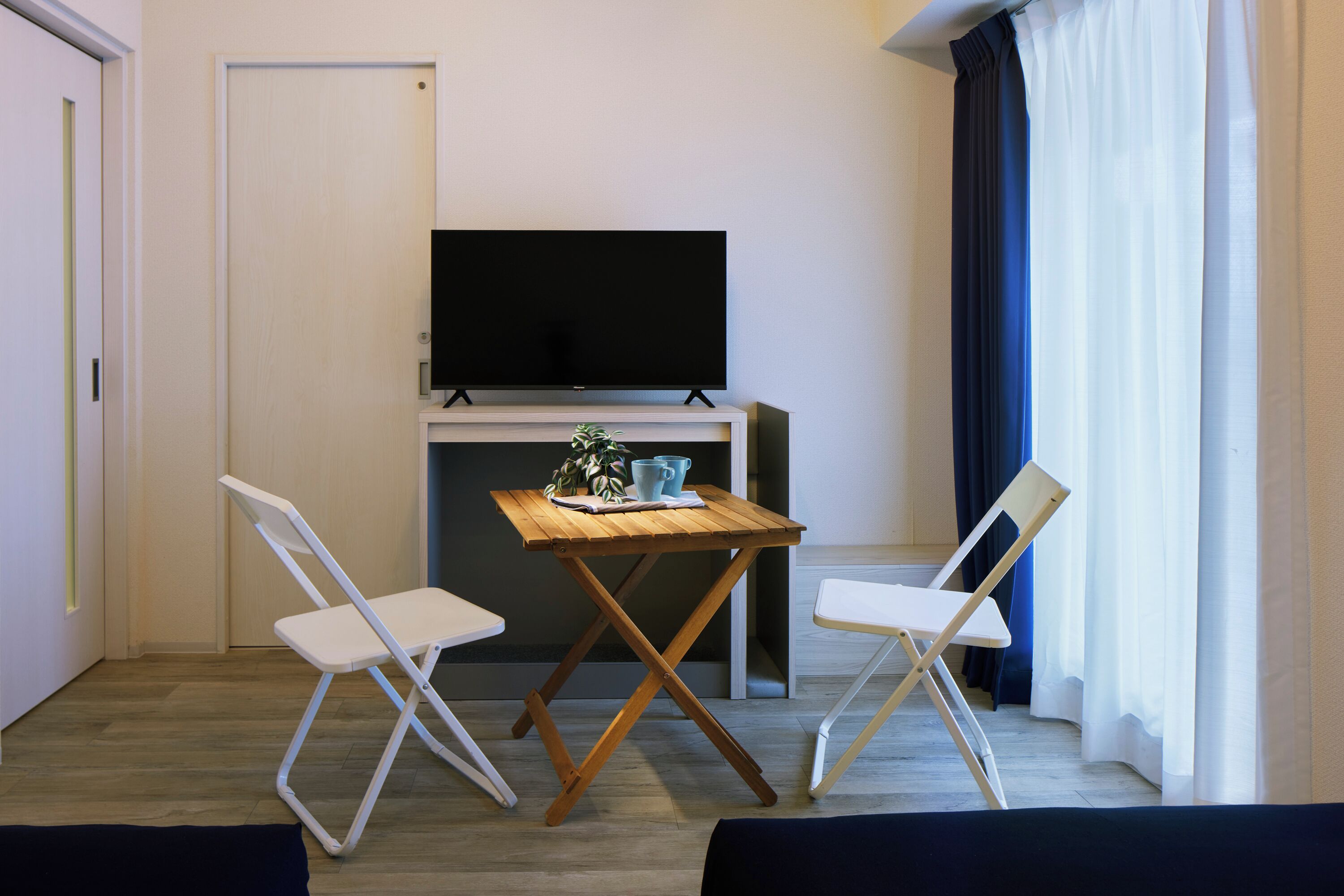 Una habitación pequeña con una televisión sobre un mueble bajo, una mesa con una planta y una taza, dos sillas blancas y una ventana con cortinas.